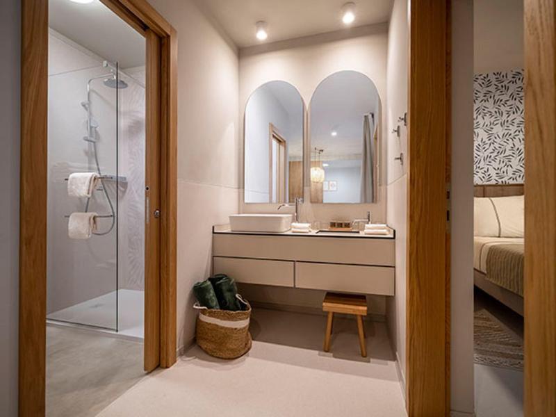 Modern bathroom area with wooden accents, double sinks, stool, and view into the bedroom.