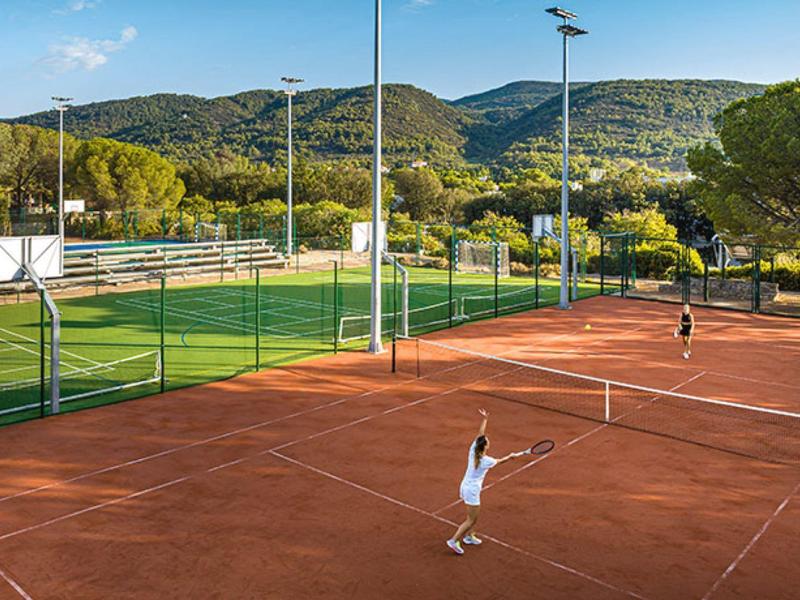 Two people playing on a red clay tennis court with mountains in the background on a sunny day.