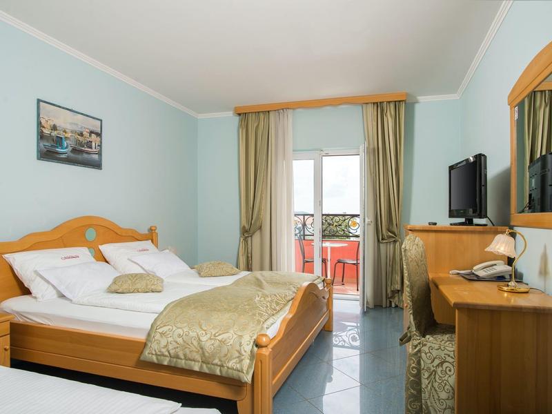 Helles Schlafzimmer mit Holzmöbeln, Doppelbett, Schreibtisch, TV und Balkon mit rotem Geländer.