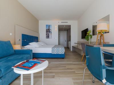 Modern hotel room with bed, sofa, table, and chairs on wooden floor.