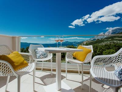 Balcony with two white chairs, table, drinks, overlooking the sea and mountains in the background.
