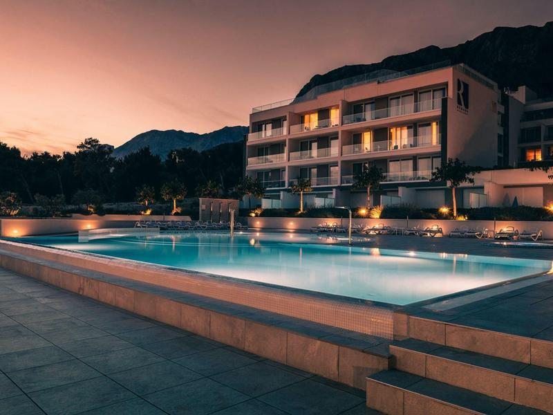 Modern hotel pool at sunset with illuminated buildings and mountains in the background.