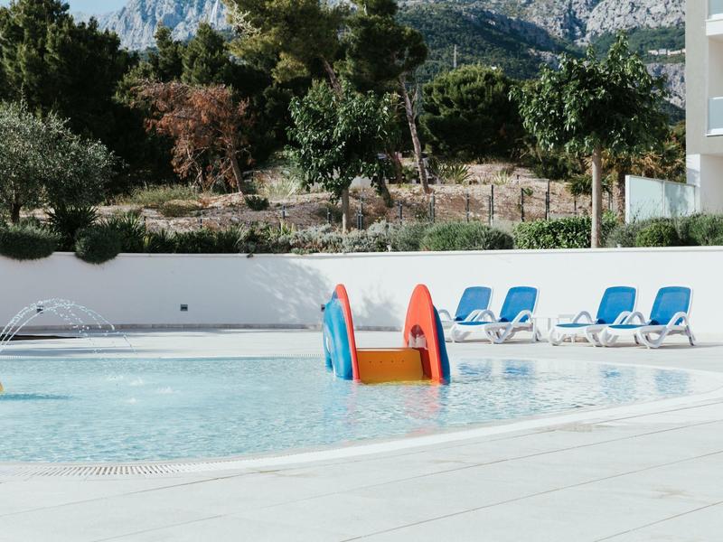 Children's wading pool with a small water slide and blue loungers next to modern hotel buildings.