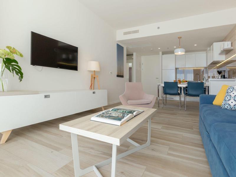 Bright modern living room with blue sofa, wall-mounted TV, and open kitchen.
