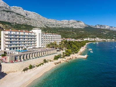 Großes Hotelgebäude am Strand mit türkisfarbenem Meer und Bergen im Hintergrund unter klarem Himmel.
