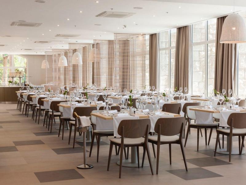 Elegancka, jasna jadalnia z białymi nakrytymi stołami i brązowymi krzesłami przy dużych oknach.