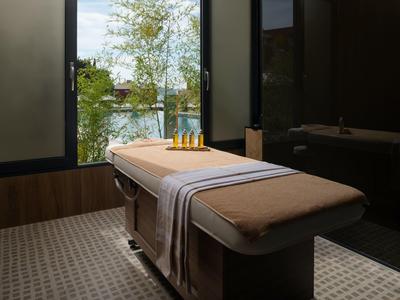 Stylová spa místnost s masážním stolem před oknem s výhledem na stromy a vodu.