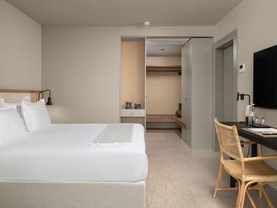 Habitación de hotel moderna con cama grande, escritorio y vestidor en tonos neutros.