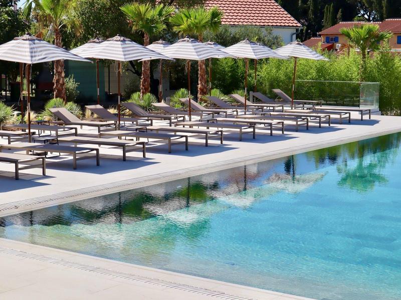 Bazénová oblast s lehátky a slunečníky vedle čisté vody a bujné zeleně.