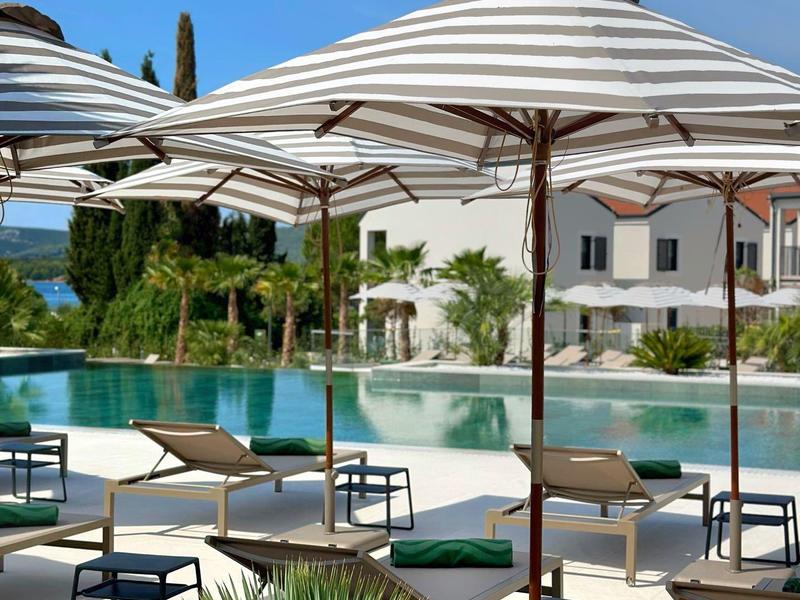 Venkovní hotelová bazénová zóna s lehátky a slunečníky pod jasnou modrou oblohou.