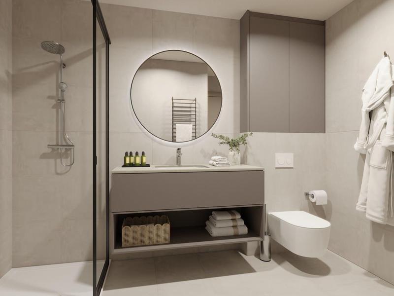 Moderní hotelová koupelna s kulatým zrcadlem, sprchovým koutem a bílými župany.