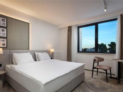 Nowoczesny pokój hotelowy z dużym łóżkiem, krzesłem, oknem i neutralnym wystrojem.