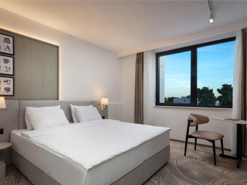 Nowoczesny pokój hotelowy z dużym łóżkiem, krzesłem, oknem i neutralnym wystrojem.