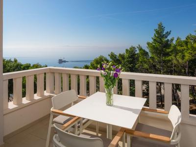 Balkon mit Tisch, Stühlen und Blumen, Blick auf Meer und bewaldete Küste bei klarem Himmel.