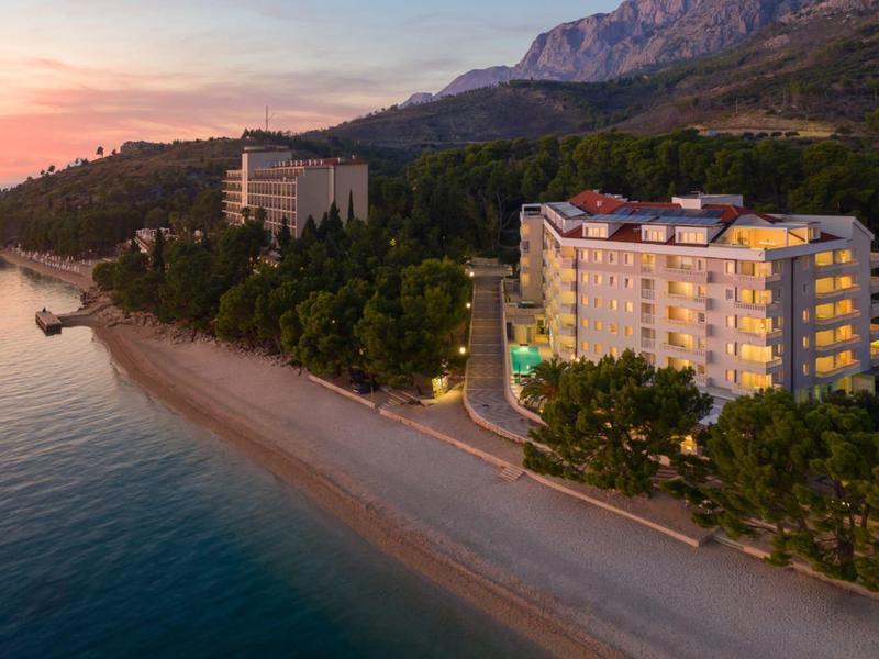 Hôtel sur une plage de sable calme avec montagnes et coucher de soleil en arrière-plan.