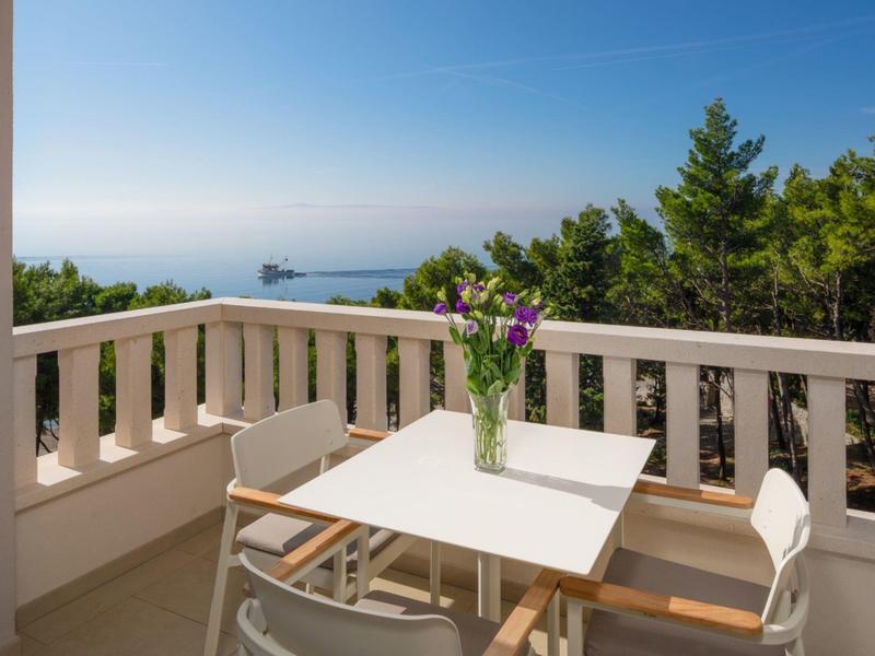 Balcon avec table blanche, chaises, fleurs, vue sur forêt et mer sous un ciel clair.