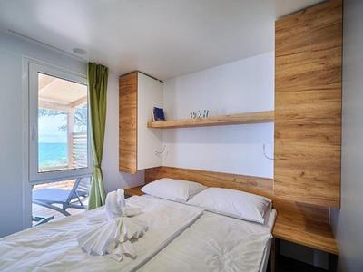 Bright hotel room with double bed, wooden shelves, and sea view through window with curtains.