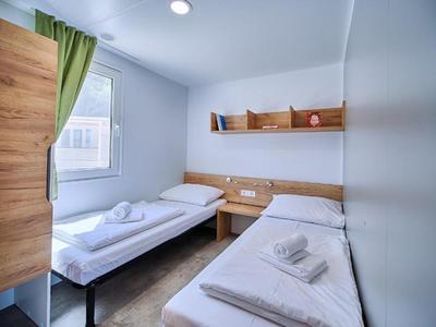 Small hotel room with two single beds, wooden shelf, and window with green curtain.