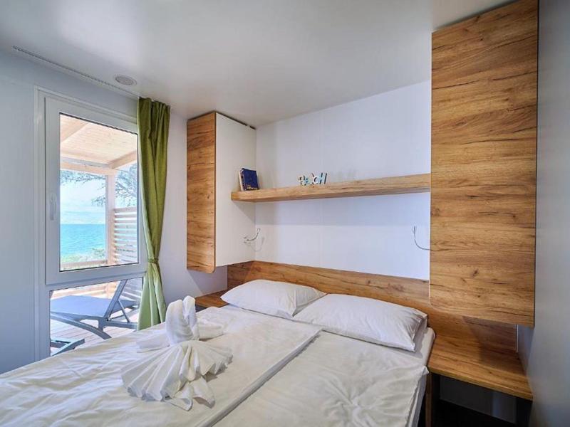 Bright hotel room with double bed, wooden shelves, and sea view through window with curtains.
