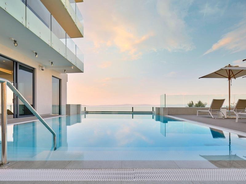 Nowoczesny basen typu infinity z leżakami i parasolem na tarasie z widokiem na morze o zachodzie słońca.