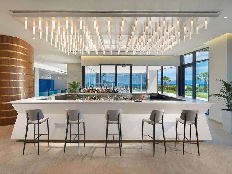 Nowoczesny bar hotelowy z oświetlonym sufitem, białymi ladami i stołkami, widok na taras i morze.
