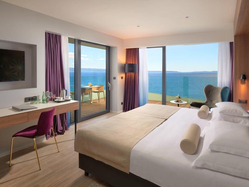 Nowoczesny pokój hotelowy z dużym łóżkiem, biurkiem i widokiem na morze przez okna od podłogi do sufitu.