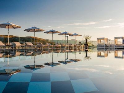 Piscina a sfioro con ombrelloni e lettini davanti a un paesaggio costiero tranquillo al tramonto.