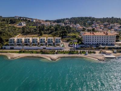 Budynki hotelowe wzdłuż wybrzeża z czystą turkusową wodą i zalesionymi wzgórzami w tle.