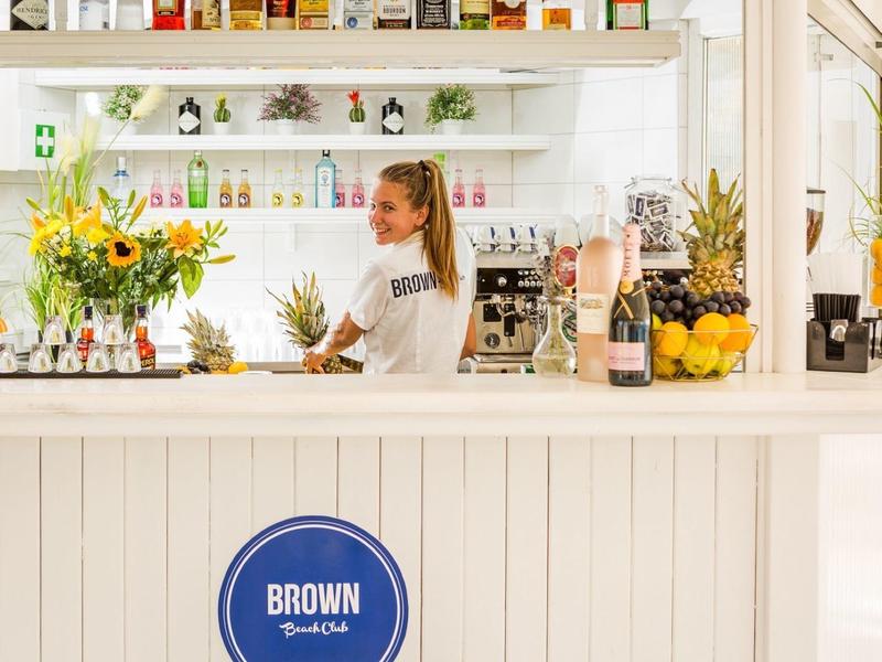 Jasno oświetlony biały bar z tropikalnymi owocami i kwiatami, kobieta za barem.