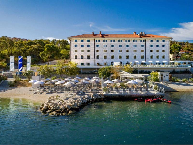 Budynek hotelu na wybrzeżu z leżakami, parasolami i molo na wodzie.