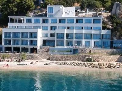 Modernes Hotel mit Glasfassade am Ufer, umgeben von Bäumen und mit Liegestühlen am Strand.