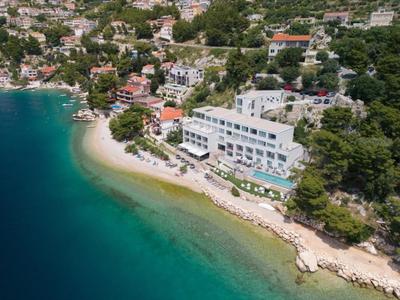 Modern hotel by a turquoise beach with green surroundings and hillside houses.