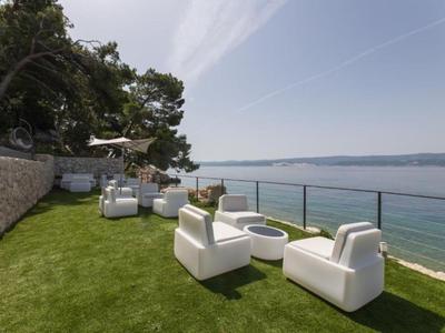 Modern seating area with white armchairs and fire pit on artificial grass by the sea.
