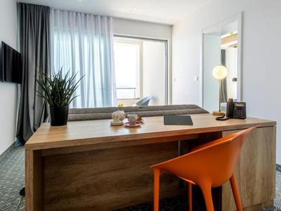 Moderner Schreibtisch mit orangefarbenem Stuhl, Pflanze und Fenster mit Vorhängen in einem Hotelzimmer.