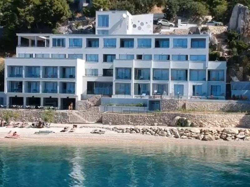 Modernes Hotel mit Glasfassade am Ufer, umgeben von Bäumen und mit Liegestühlen am Strand.