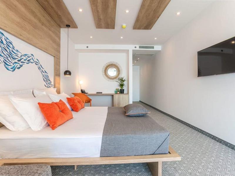 Heller Hotelzimmer mit Doppelbett, grauer Bettwäsche, orangenen Kissen, Holzdecke und Wand-TV.