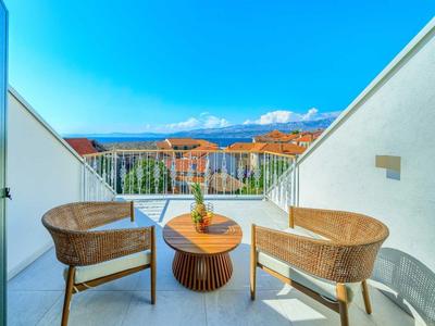 Balkon mit zwei Korbstühlen, Holztisch und Blick auf das Meer und die Berge bei klarem Himmel.