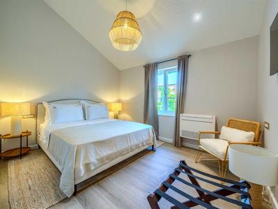 Hell eingerichtetes Schlafzimmer mit Doppelbett, Korbstuhl und Fenster mit Vorhängen.