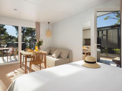 Helles Hotelzimmer mit Bett, Sofa, Esstisch und Zugang zur Terrasse mit Gartenblick.