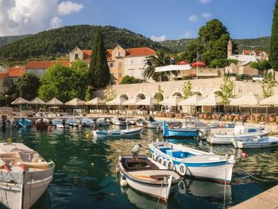 Kleine Boote schwimmen in ruhigem Wasser vor einer Hotelanlage mit Sonnenschirmen und Bergen im Hintergrund.