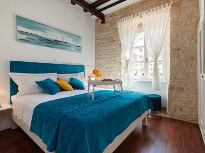 Gemütliches Hotelzimmer mit Doppelbett, blauen Bettdecken, Fenster mit weißem Vorhang und Holzbalkendecke.