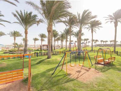 Spielplatz mit Schaukeln und Rutschen unter Palmen auf einer Hotelwiese bei Sonnenlicht.