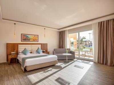 Helles Hotelzimmer mit Doppelbett, Sofa und Balkon mit Blick auf Poolbereich.