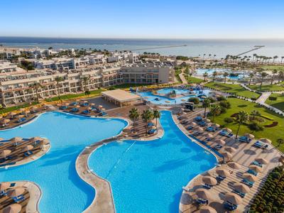 Großer Hotelpool mit Sonnenliegen, Palmen und Blick auf das Meer bei klarem Himmel.