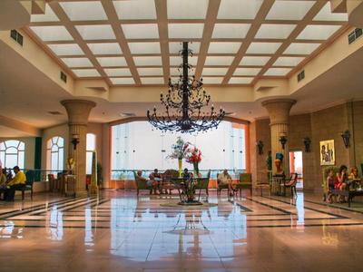 Großer Hotellobby mit Kronleuchter, Sitzbereich und hohen Säulen unter hellem Glasdach.