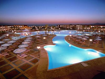 Großer beleuchteter Pool mit Liegestühlen und Sonnenschirmen in einem Resort bei Sonnenuntergang.