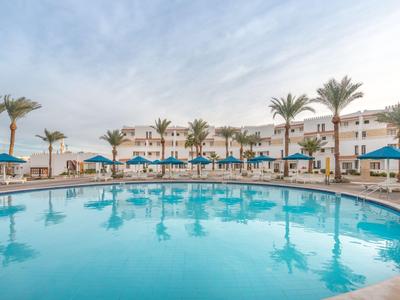 Großer, runder Hotelpool mit blauen Sonnenschirmen und Palmen bei klarem Himmel.