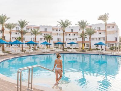 Frau steigt in einen großen, runden Pool vor einem weißen Hotel mit blauen Sonnenschirmen.