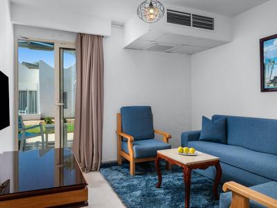 Moderní hotelový obývací prostor s modrou pohovkou, křeslem, kobercem a přístupem na balkon.