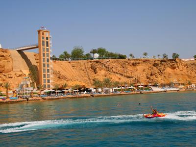 Küste mit rotem Felsen, Turm, Hotel und einem Jetboot auf türkisfarbenem Wasser.